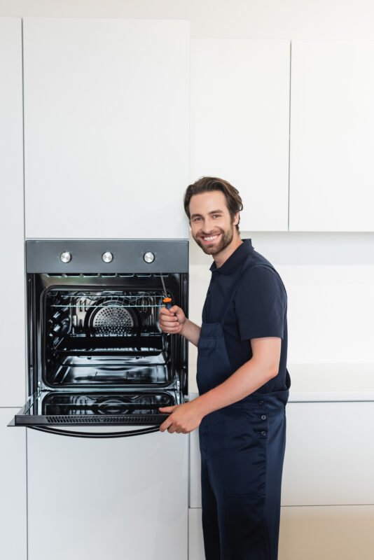 happy repairman with screwdriver smiling at camera near electric oven