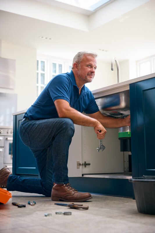 Male Plumber Fixing Waste Disposal Unit In Domestic Kitchen Sink