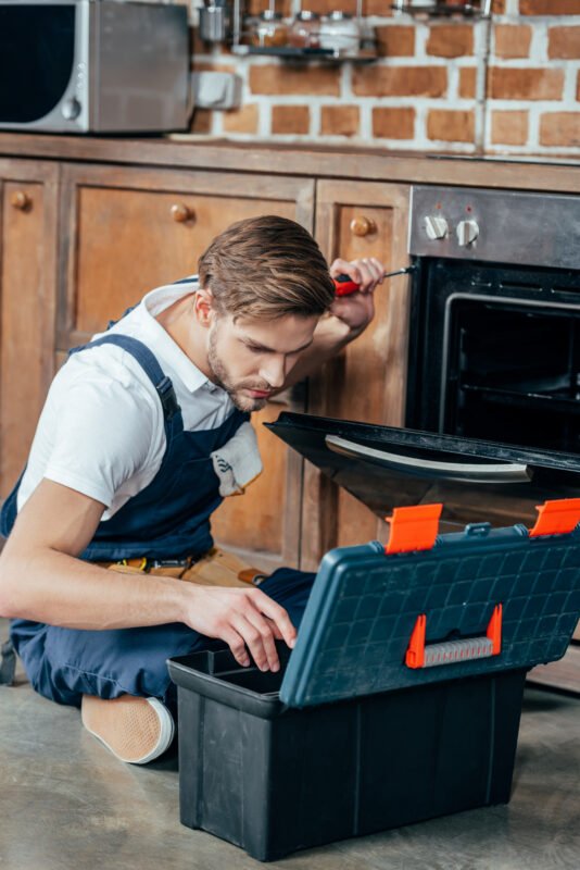 repairman looking at toolbox while fixing oven with screwdriver