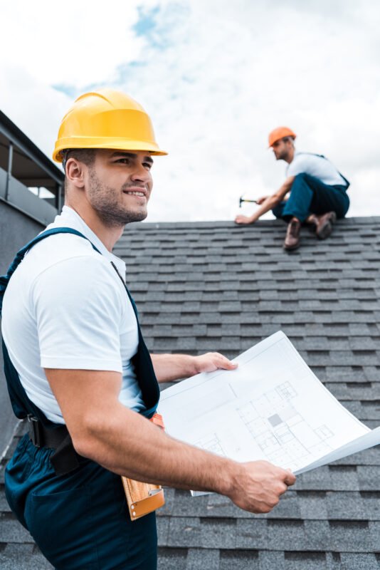 Builder in helmet holding blueprint near colleague repairing roof